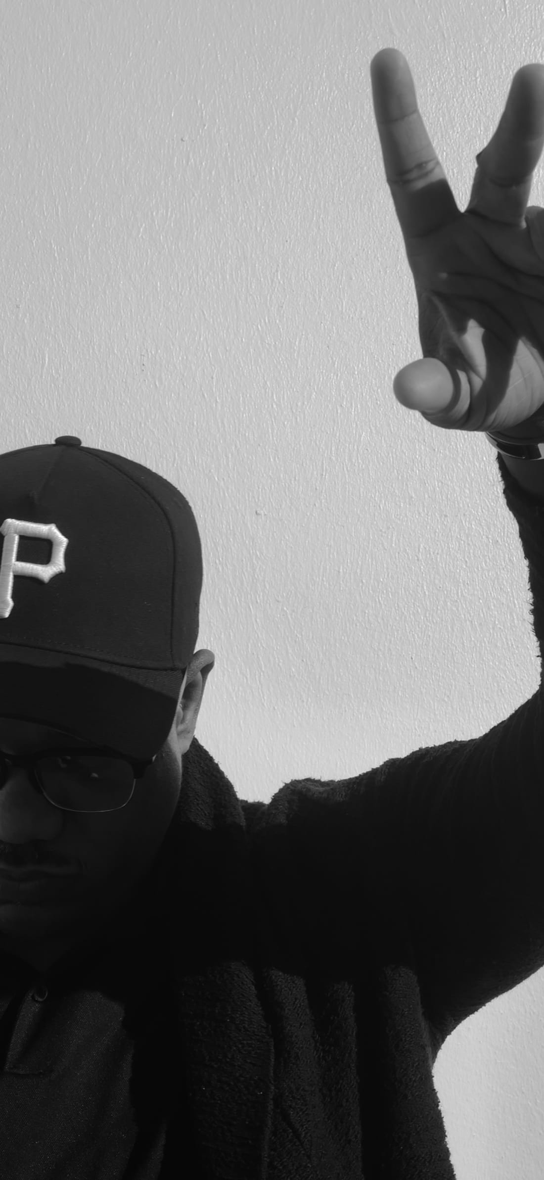 Lechon Kirb in a black Pittsburgh ball cap against a pale wall, one hand raised, photographed in black and white