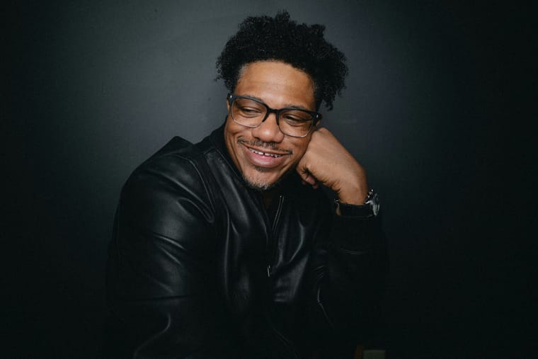 Lechon Kirb in a black leather jacket and glasses, chin resting on his hand, smiling