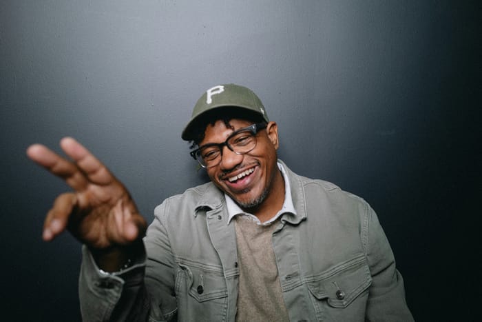 Lechon Kirb laughing in a green Pittsburgh ball cap and olive jacket, one hand raised toward the camera